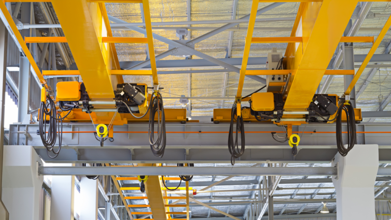 Vue en contre-plongée de deux ponts roulants industriels jaunes installés sur des rails au plafond d'une usine. On distingue nettement les mécanismes de levage, les câbles et la structure métallique du bâtiment.