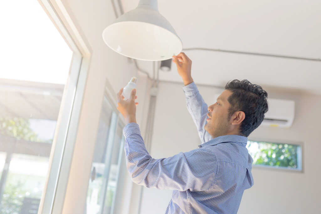 Un homme en chemise bleue installe ou remplace une ampoule LED dans un luminaire suspendu blanc, à côté d'une grande fenêtre lumineuse.