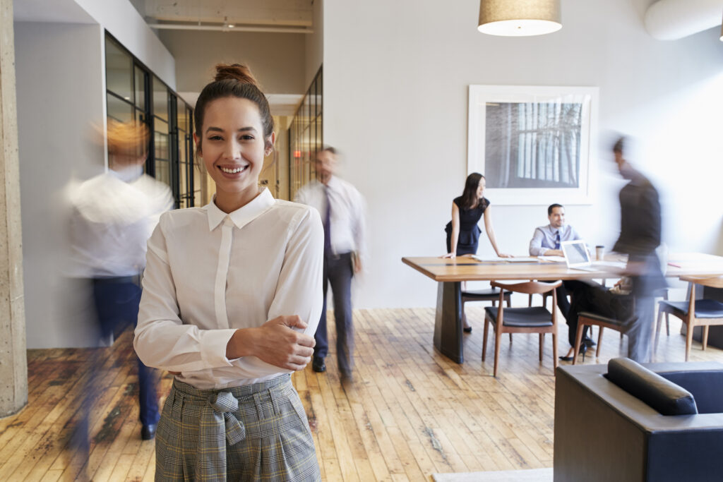 Une jeune femme souriante, bras croisés, au premier plan d'un bureau moderne et lumineux. En arrière-plan, ses collègues sont en mouvement ou travaillent autour d'une grande table en bois, illustrant une ambiance de travail dynamique.
