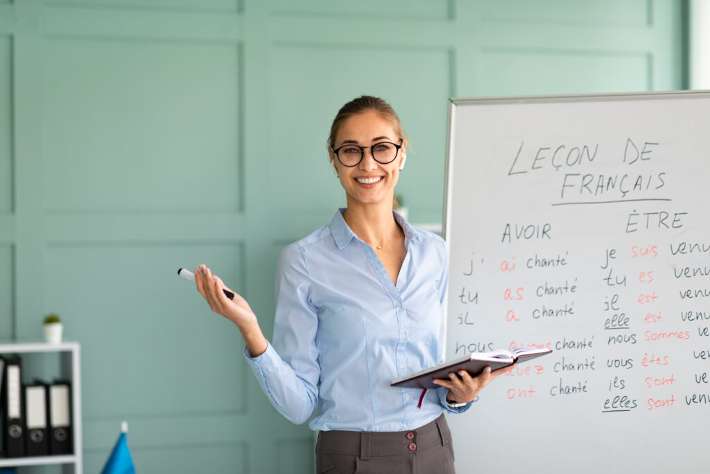 Une jeune femme souriante, portant des lunettes et une chemise bleue, se tient devant un tableau blanc. Elle tient un stylo dans une main et un carnet ouvert dans l'autre. Le tableau affiche une leçon de français intitulée « LEÇON DE FRANÇAIS », avec les conjugaisons des verbes « AVOIR » et « ÊTRE » écrites de manière organisée. L'arrière-plan montre un mur vert pâle et une étagère, créant une ambiance de salle de classe ou de bureau.