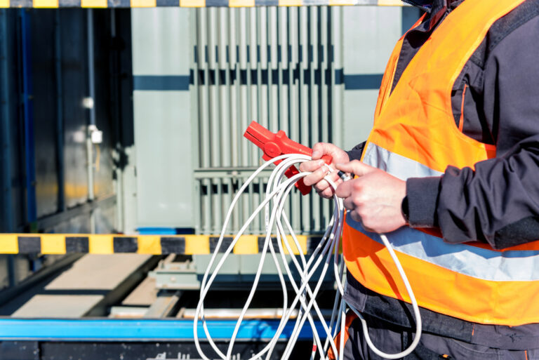 Un technicien de maintenance en gilet de sécurité tenant des câbles et une pince de mesure devant un transformateur électrique.
