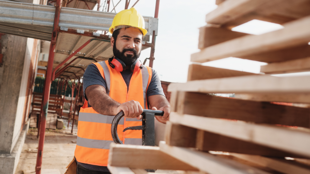 un ouvrier du bâtiment barbu, portant un casque de sécurité jaune et un gilet orange, déplace une pile de palettes en bois à l'aide d'un transpalette manuel sur un chantier.