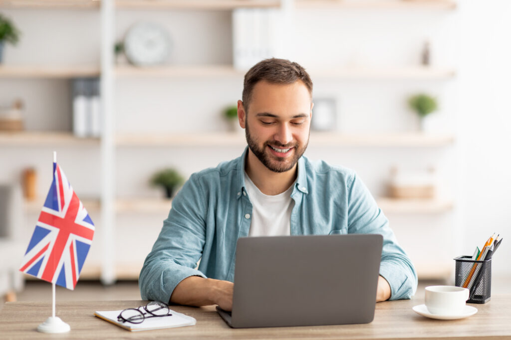 Un homme souriant à barbe courte, vêtu d'une chemise en jean bleu clair, travaille sur un ordinateur portable gris à son bureau. Sur la table, on aperçoit un petit drapeau du Royaume-Uni, une paire de lunettes posée sur un carnet, une tasse de café blanche et un pot à crayons. Le fond montre des étagères de bureau claires et légèrement floues avec quelques plantes vertes.
