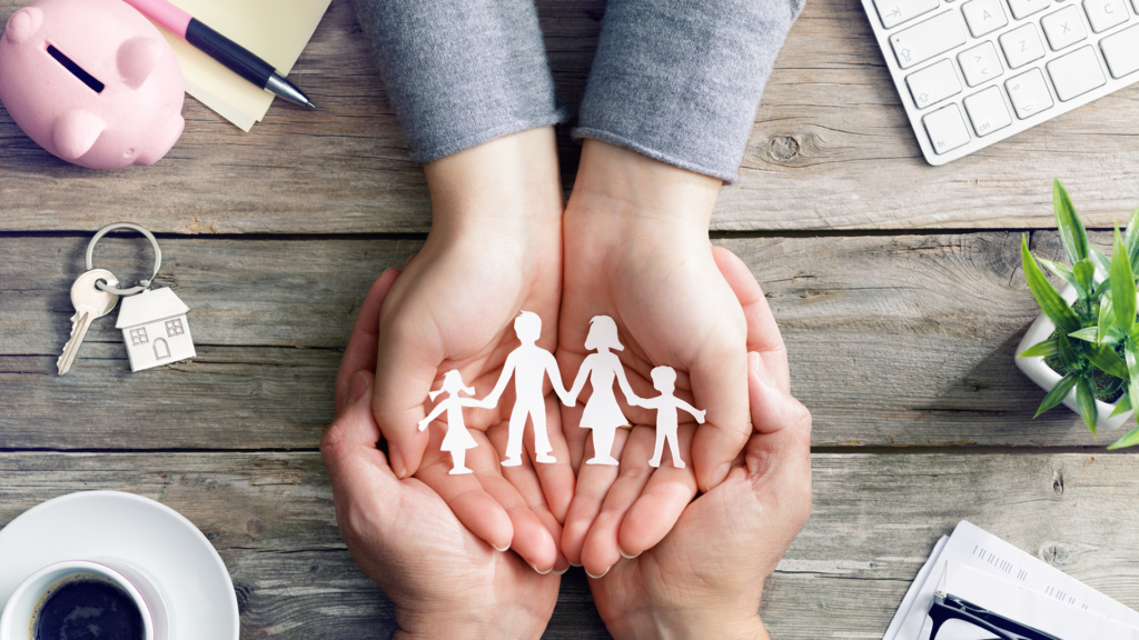 Deux mains jointes en coupe protégeant une découpe en papier représentant une famille de quatre personnes sur un bureau en bois.