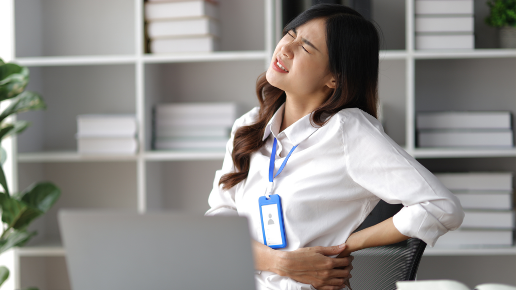 une personne assise devant un ordinateur et se tenant le bas du dos avec une expression de douleur, ce qui semble être une représentation de troubles musculosquelettiques
