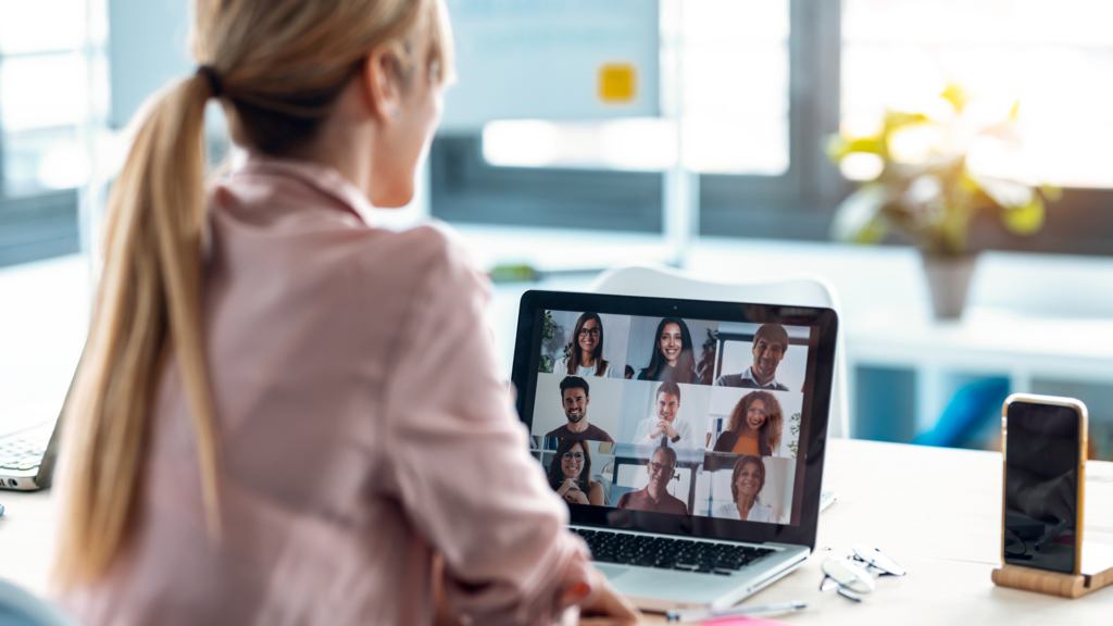 Une femme aux cheveux blonds attachés en queue de cheval travaille à son bureau et participe à une réunion virtuelle affichant plusieurs collègues sur l'écran de son ordinateur.