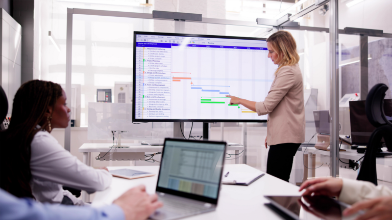 Une femme présente un diagramme de Gantt sur un grand écran interactif lors d'une réunion de projet. Ses collègues, assis autour d'une table avec des ordinateurs portables, suivent attentivement la planification dans un bureau moderne et lumineux