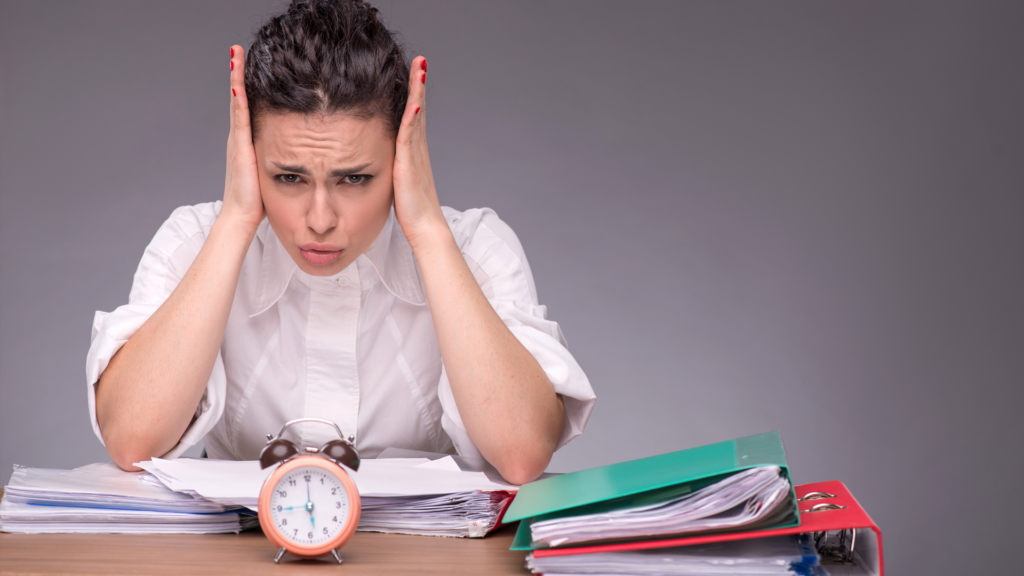 Une femme brune, l'air stressé et fatigué, est assise à son bureau. Elle se couvre les oreilles avec ses mains, comme pour bloquer le bruit ou la pression. Devant elle se trouvent des piles de dossiers, des classeurs colorés et un réveil rose à double cloche qui semble symboliser l'urgence ou une échéance proche.